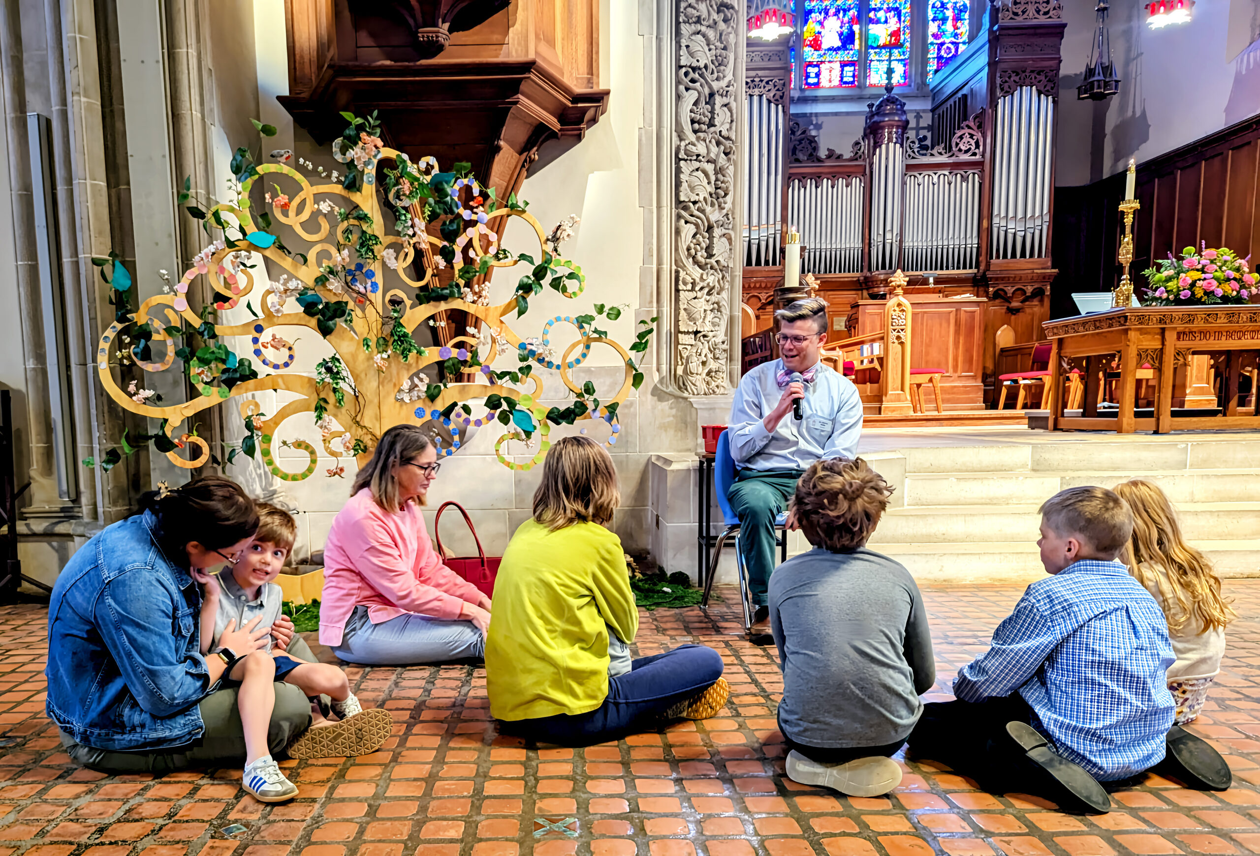 Children and adults gather on the sanctuary floor at House of Hope, listening to a speaker beside a colorful tree display.