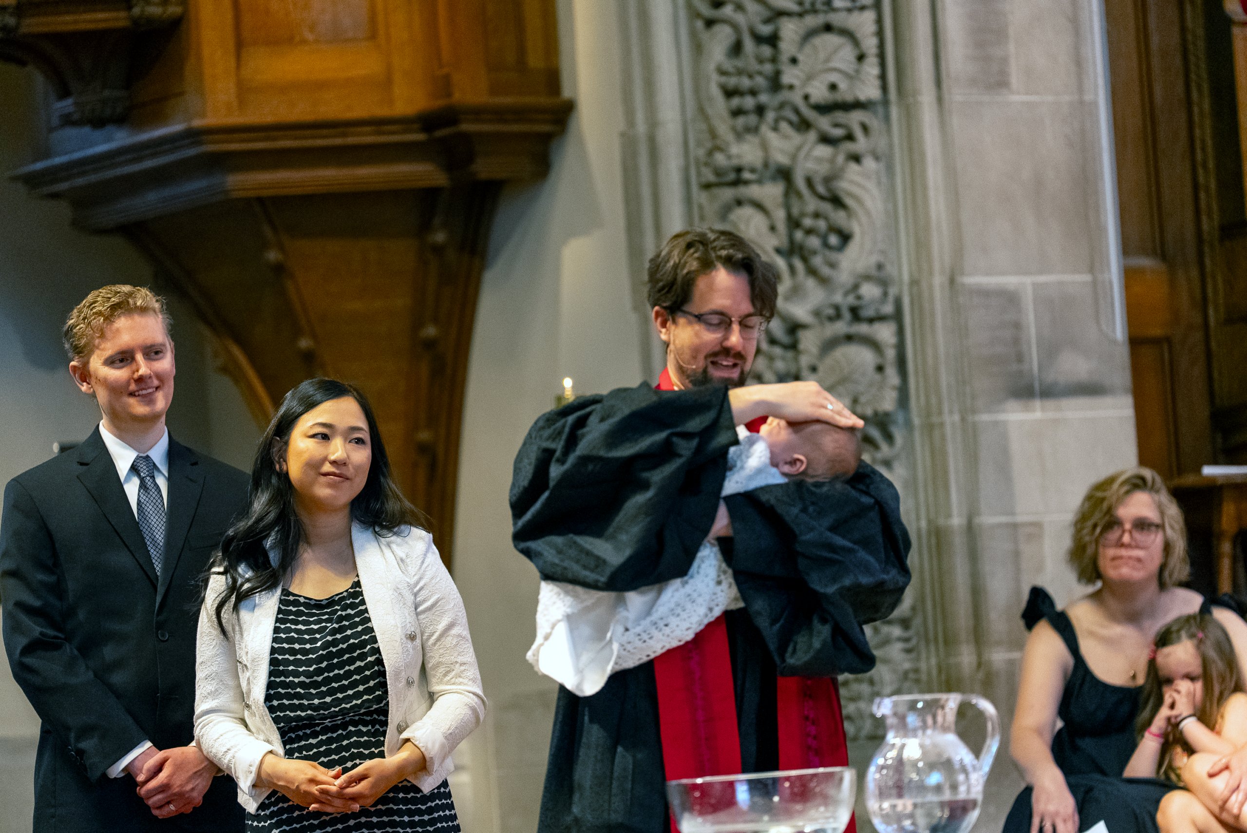 A minister baptizes an infant beside the baptismal font, with family members nearby.